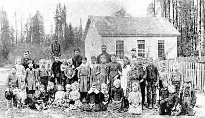 Teachers and students, small one-story frame building, slat fence, tall evergreens.