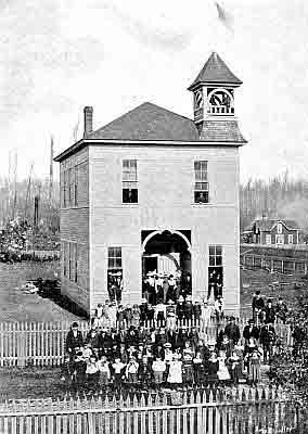 Tall two story frame building, bell tower; people of all ages on steps and risers; picket fences.